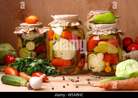 Un assortimento di verdure sottaceto in vasi: cetrioli e pomodori, cavolo, zucchine e peperoni con aglio, aneto e foglie di alloro in vasi su un di legno backgroun Foto Stock