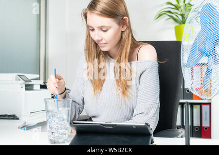 Una giovane donna in ufficio non si raffredda con un bicchiere di acqua pieno di cubetti di ghiaccio Foto Stock