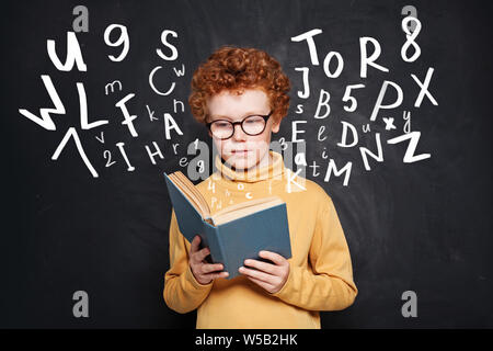 Curioso ragazzino con gli occhiali per leggere un libro contro la lavagna Foto Stock