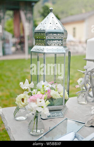 Primo piano di un grande vetro lanterna decorativo decorato con fiori e piccoli vasi con fiori su uno sfondo sfocato di un prato verde, selettivo foc Foto Stock