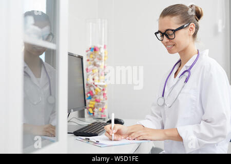 Vista laterale ritratto di donna sorridente medico iscritto negli appunti mentre è seduto alla scrivania in ufficio, spazio di copia Foto Stock