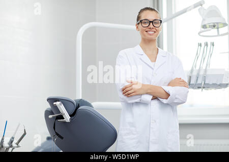 Ritratto di giovane donna medico che indossano occhiali sorridente alla telecamera mentre posa con poltrona odontoiatrica, spazio di copia Foto Stock