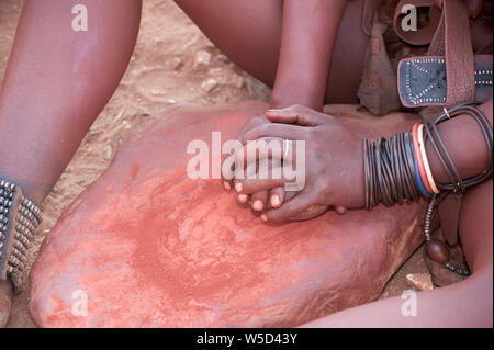 Donna Himba schiaccia l'argilla rossa, questo viene poi utilizzato per rivestire i loro capelli. Fotografato in un villaggio Himba, Epupa Falls, Kaokoland, Namibia, Africa Foto Stock