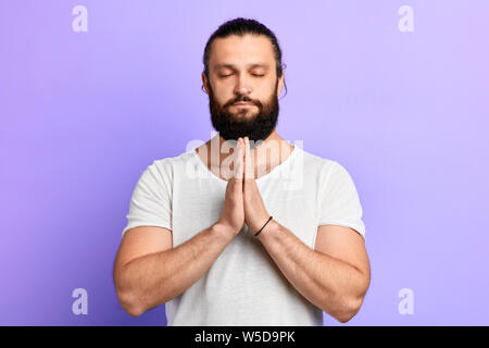 Sprituality e calma uomo tiene le mani insieme con gli occhi chiusi, isolate su backgrouund blu. I religiosi giovane augura salute per la sua famiglia in stu Foto Stock