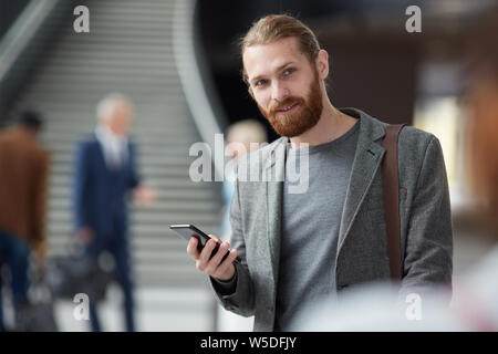 Ritratto di positivo rosso-barbuto ragazzo in giacca grigia in piedi nella lobby e utilizzando gadget Foto Stock