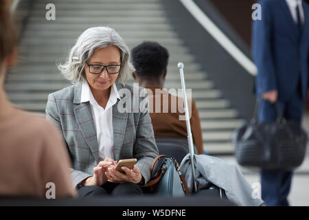 Contenuto anziani donna asiatica con i capelli grigi che indossa occhiali seduto in area di attesa di airport e utilizzi mobile app sul gadget Foto Stock