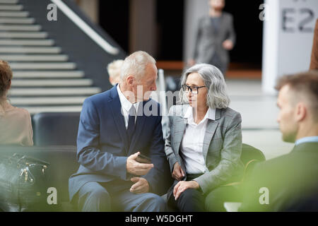 Grave uomo maturo in abito formale seduti sul divano in aeroporto e spiegando meta di viaggio di affari al collega asiatica Foto Stock