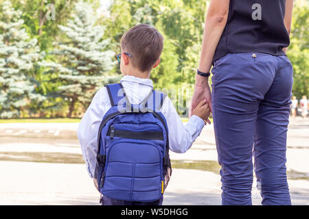 Il ragazzo è un primo-grader in bicchieri con una valigetta blu tenendo la sua mano madre. Si torna a scuola Foto Stock