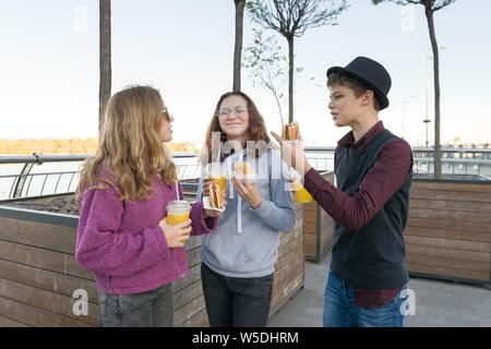 Gli adolescenti mangiare cibo di strada, amici un ragazzo e due ragazze su una strada di città con hamburger e succo di arancia. Sullo sfondo della città, ora d'oro Foto Stock