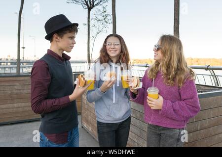 Gli adolescenti mangiare cibo di strada, amici un ragazzo e due ragazze su una strada di città con hamburger e succo di arancia. Sullo sfondo della città, ora d'oro Foto Stock