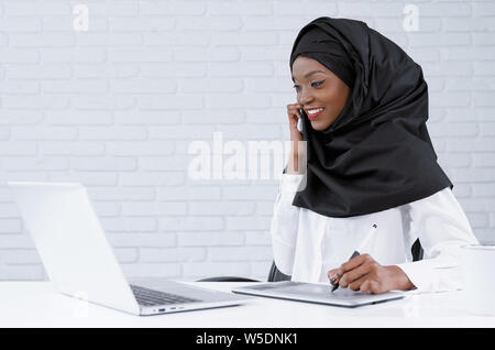 Bella donna musulmana parla al telefono, guardando il laptop, sorridente. Affascinante e positivo ufficio africano Lavoratore che indossa in nero hijab e abiti bianchi seduta a tavola con il computer. Foto Stock