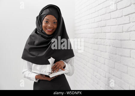 Piuttosto, allegro africano donna musulmana in piedi vicino al bianco muro di mattoni, tenendo notebook. Bella, positivo lavoratore di ufficio che indossa in nero hijab, camicia bianca guardando la telecamera, sorridente. Foto Stock