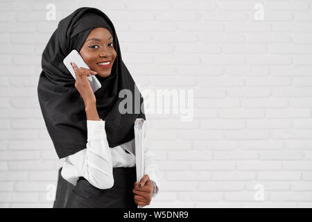 Elegante e con stile donna musulmana che indossa in nero hijab holding cartella bianca con i documenti e il telefono cellulare. Ufficio africano ragazza con sorriso perfetto guardando la telecamera, in posa. Foto Stock