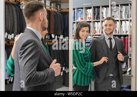 Client sorridente in maglietta, gilet, giacca e cravatta cercando in corrispondenza dello specchio di boutique. Assistente ragazza di aiutare e di scelta di abbigliamento per uomo giovane. Moda e stile per la riuscita dell'uomo. Foto Stock