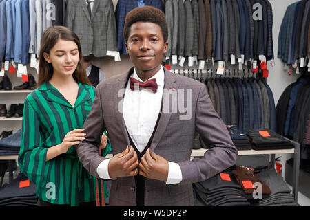 Bello l'uomo africano con capelli ricci in camicia bianca, gilet, giacca grigio e rosso il filtro bow tie in posa. Client di giovani montaggio elegante abito, guardando la fotocamera. Ragazza, shop consultant in piedi vicino all'uomo. Foto Stock