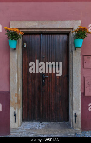 Storico cancello in legno di una casa residenziale con vasi da fiori. Rosso e marrone a parete con lastre di granito sul palco. chiuso la porta di legno con raccordo in metallo Foto Stock