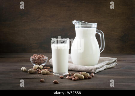 Fresco latte di arachidi in vetro e bricco scuro su un tavolo di legno. Vegano sano di mangiare. Stile rustico. Foto Stock
