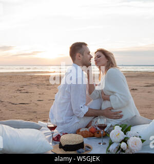 Picnic al tramonto. Un uomo e una donna incinta di sedersi su un copriletto, fra i frutti e i cuscini, sulla spiaggia, avvolgente, baciare. Foto Stock