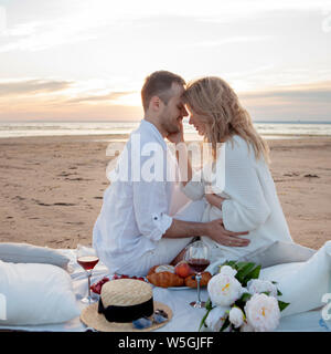 Picnic al tramonto. Un uomo e una donna incinta di sedersi su un copriletto, fra i frutti e i cuscini, sulla spiaggia, avvolgente, baciare. Foto Stock