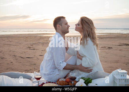 Picnic al tramonto. Un uomo e una donna incinta di sedersi su un copriletto, fra i frutti e i cuscini, sulla spiaggia, avvolgente, baciare. Foto Stock