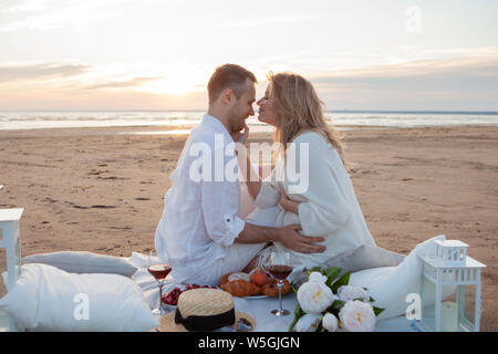 Picnic al tramonto. Un uomo e una donna incinta di sedersi su un copriletto, fra i frutti e i cuscini, sulla spiaggia, avvolgente, baciare. Foto Stock