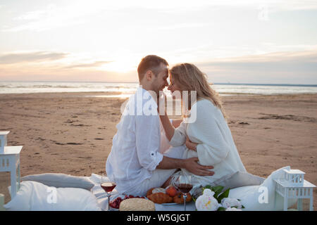 Picnic al tramonto. Un uomo e una donna incinta di sedersi su un copriletto, fra i frutti e i cuscini, sulla spiaggia, avvolgente, baciare. Foto Stock