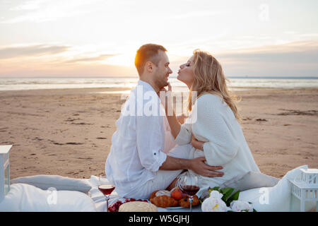 Picnic al tramonto. Un uomo e una donna incinta di sedersi su un copriletto, fra i frutti e i cuscini, sulla spiaggia, avvolgente, baciare. Foto Stock