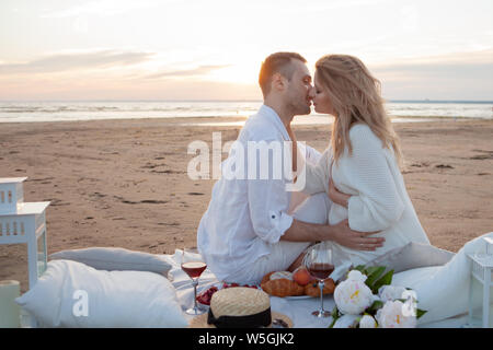 Picnic al tramonto. Un uomo e una donna incinta di sedersi su un copriletto, fra i frutti e i cuscini, sulla spiaggia, avvolgente, baciare. Foto Stock