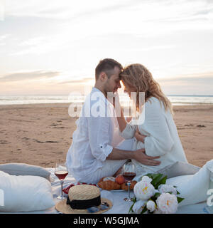 Picnic al tramonto. Un uomo e una donna incinta di sedersi su un copriletto, fra i frutti e i cuscini, sulla spiaggia, avvolgente, baciare. Foto Stock