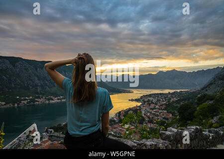 Montenegro, giovane e bella bionda signora seduta sulla pietra antica parete sopra kotor bay city nel fantastico arancione tramonto crepuscolo sulle calde vacanze estive Foto Stock