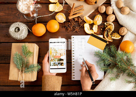 Mani femminili tenendo lo smartphone e rendendo lista dei regali di Natale Foto Stock