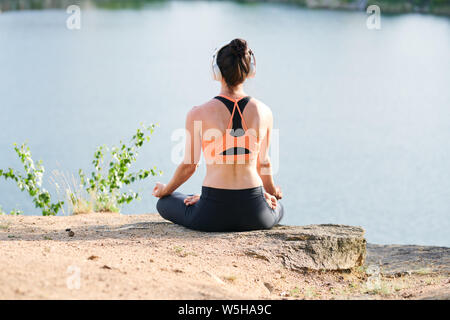 Vista posteriore della giovane donna in cuffie wireless seduti nella posizione del loto Foto Stock