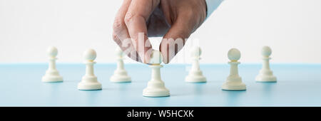 Ampia vista immagine dei maschi di mano che tiene la prima di molte pedina scacchi pezzi posti sulla scrivania blu in una immagine concettuale. Foto Stock