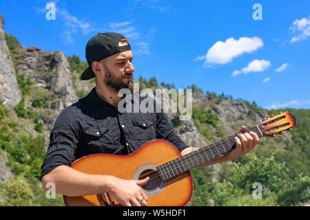 Giovani hipster barbuto guy, uomo in un cappuccio suona la chitarra contro lo sfondo di un luminoso paesaggio estivo, cielo blu e montagne Foto Stock