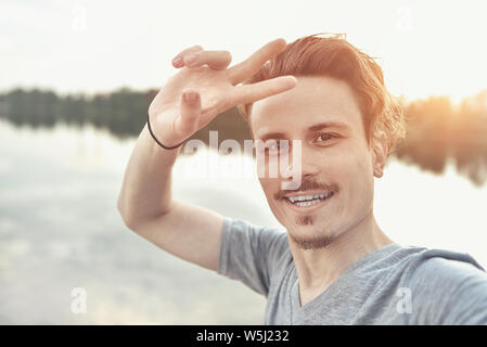 Ritratto di giovane bello ed elegante ragazzo sorridente rende selfie contro il lago. bellissima natura Foto Stock