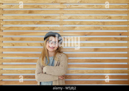 Carino sorridente bambina con nero indossando occhiali cappuccio su sfondo di legno. L'educazione, la scuola dell'infanzia, le persone e il concetto di visione Foto Stock