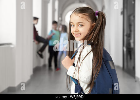 Graziosa bambina con divertenti acconciatura guardando sopra la spalla alla fotocamera e sorridente, in piedi sul corridoio nella scuola. Ragazza con zaino andare in aula a lezione. Bambini in background. Foto Stock