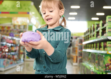 Carino e bella ragazza tenendo in mano viola sabbia cinetica, ponendo in telecamera. Piccolo bambino che gioca nella riproduzione di stanza in negozio del centro commerciale. Ripiani con i giocattoli sullo sfondo. Foto Stock