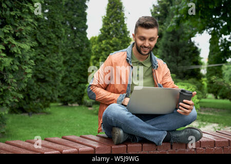 Bello, elegante uomo al lavoro su banco di legno in giardino con il laptop. Studente seduto con le gambe incrociate, tenendo la carta di tazza di caffè, cercando su laptop, sorridente. Foto Stock