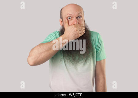 Sarò tranquilla. Ritratto di scioccato di mezza età uomo calvo con barba lunga in verde chiaro t-shirt in piedi, che copre la sua bocca, guardando con occhi grandi. Foto Stock