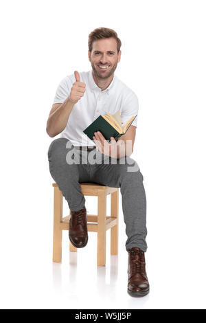 Sorridente uomo seduto a leggere un libro e rende il segno ok su sfondo bianco Foto Stock