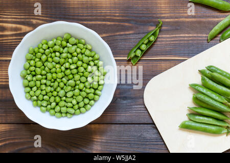 Ciotola bianco con verde di piselli pelati interno scuro su un tavolo di legno. Tutto baccelli di piselli sul tavolo da cucina e aperta singola cialda vicino a. Vista superiore dello sfondo. Persone fisiche o Foto Stock