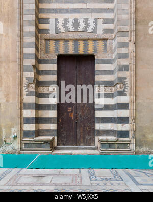 Invecchiato in legno porta circondato da strisce in bianco e nero, decorazioni in marmo e pietra parete presso il cortile del sultano moschea Hassan al Cairo, Egitto Foto Stock