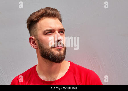 Ritratto di giovane attraente barbuto uomo caucasico con sguardo serio in T-shirt rossa su uno sfondo grigio. Foto Stock