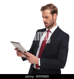 Vista laterale del businessman scioccato che indossa cravatta rossa e la marina tuta la lettura dal suo pad mentre in piedi su sfondo bianco, ritratto foto Foto Stock