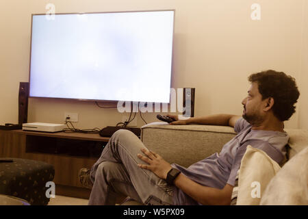 L'uomo guarda la TV di casa in soggiorno. Uomo asiatico in blu T shirt seduto su un divano a guardare la televisione sul divano. vista laterale. Tempo libero, tecnologia, Foto Stock