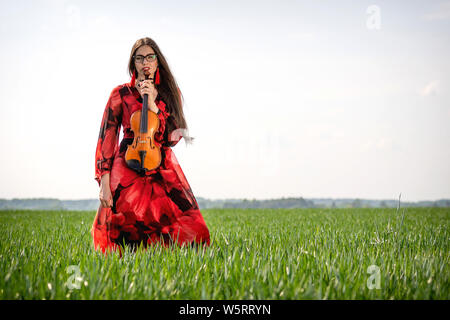 Giovane donna in abito rosso con violino in prato verde. Foto Stock