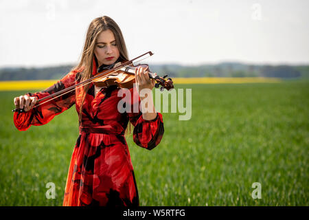 Giovane donna in abito rosso suona il violino in prato verde. Foto Stock