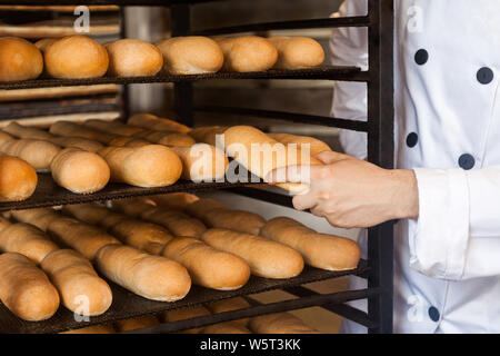 Primo piano delle mani di chef professionale uomo in bianco uniforme in piedi vicino a ripiani in pieno con pane fresco e tirando il vassoio di cottura per il controllo di qualità, ho Foto Stock