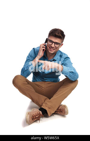 Seduto uomo informale parlando al telefono puntare il dito a lato su sfondo bianco Foto Stock